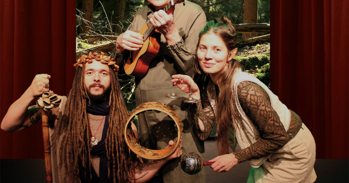Drie mensen poseren in een bosomgeving, met muziekinstrumenten.