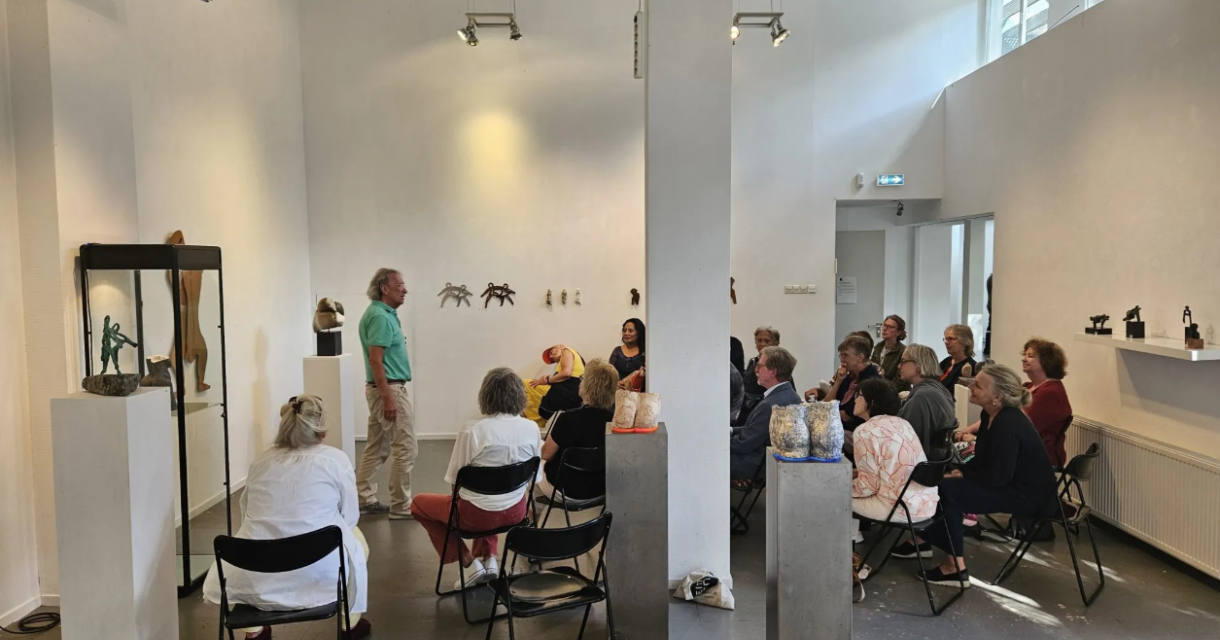 Groep mensen zit op klapstoelen in een witte galerie, luistert naar spreker tussen keramische kunstobjecten