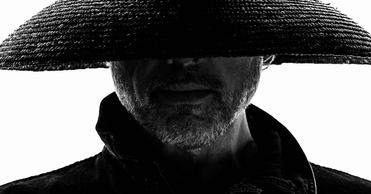 Man met baard in silhouet, draagt een brede rieten hoed die zijn ogen bedekt