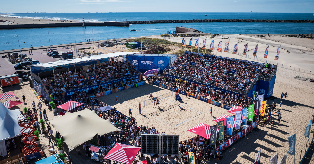 Een druk strandvolleybaltoernooi met publiek en een blauwe lucht.