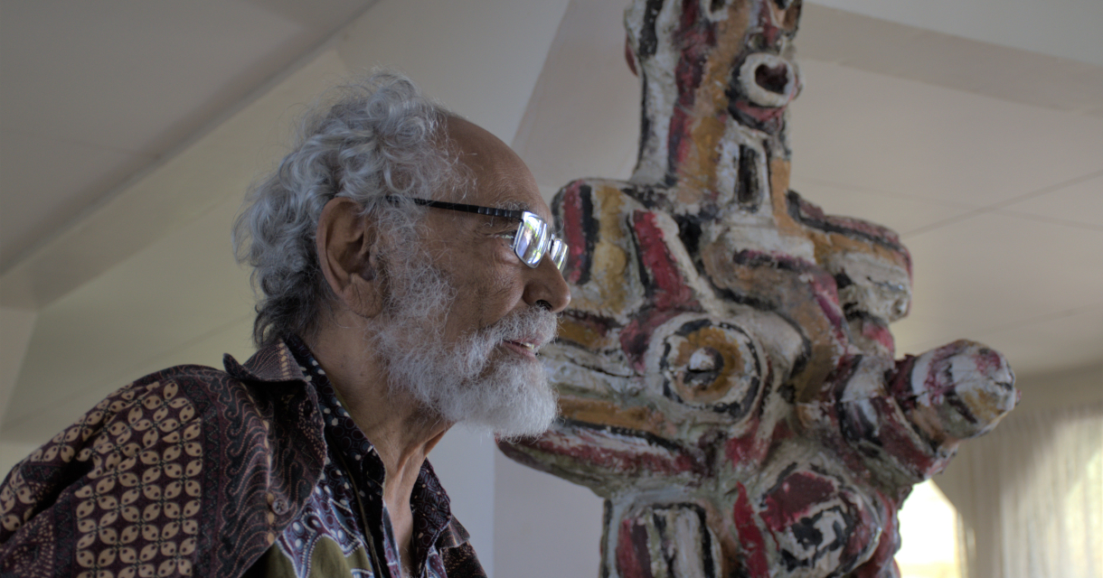 Een man met grijs haar en een baard poseert naast een kleurrijk beeldhouwwerk.
