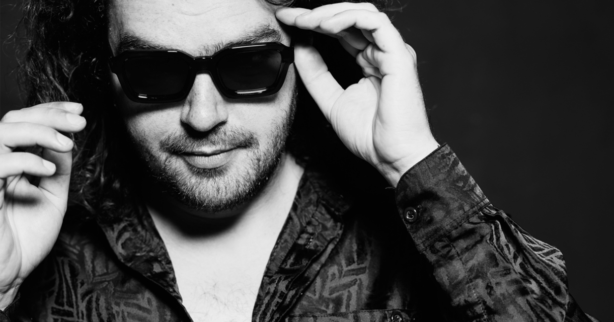 Een man met krullend haar en een zonnebril poseert.