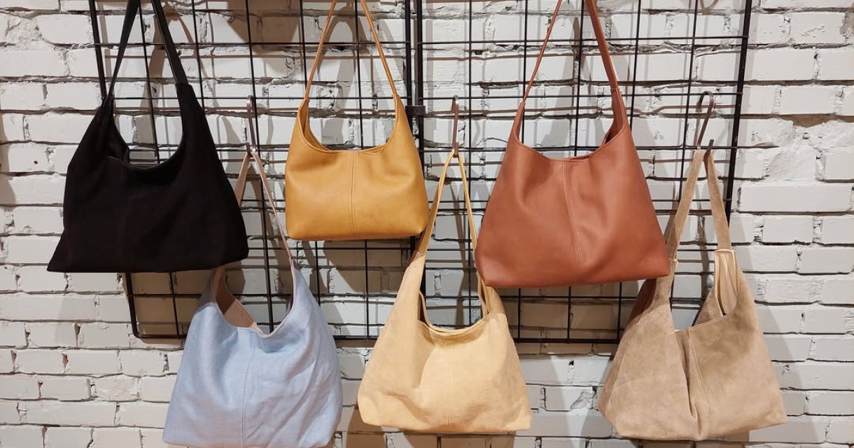 Zes schoudertassen in neutrale en aardetinten aan een zwart metalen rek tegen een witte bakstenen muur. Zes schoudertassen in neutrale en aardetinten aan een zwart metalen rek tegen een witte bakstenen muur.