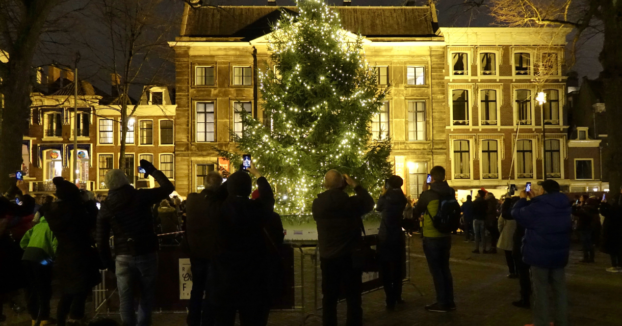 een groep mensen die 's avonds rond een verlichte kerstboom verzameld zijn op een plein. De kerstboom is versierd met lichtjes, en op de achtergrond zijn verlichte historische gebouwen te zien