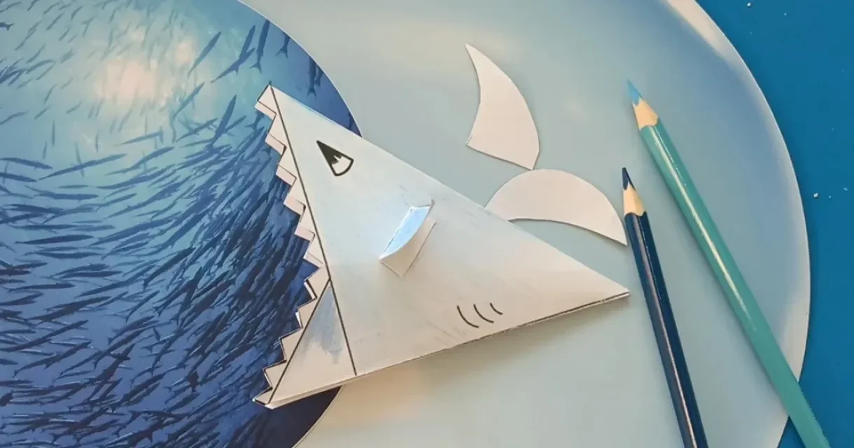 Een papieren haai ligt op een bord met golven en potloden.