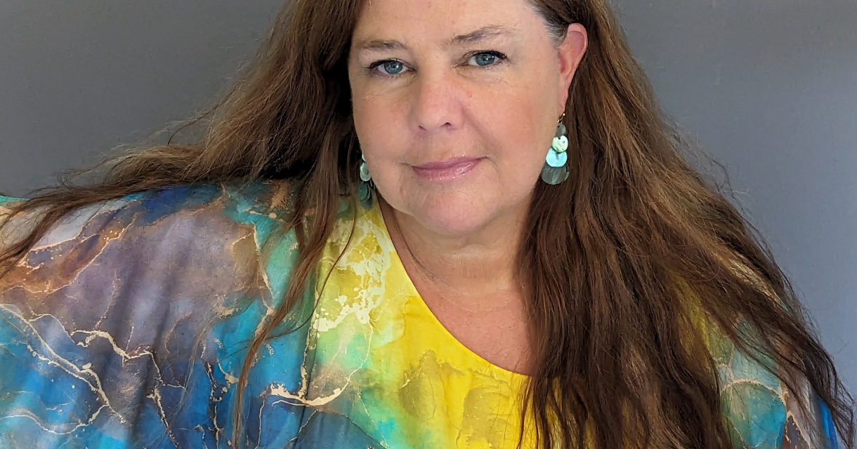 Vrouw met lang bruin haar en blauw-geel blouse poseert.