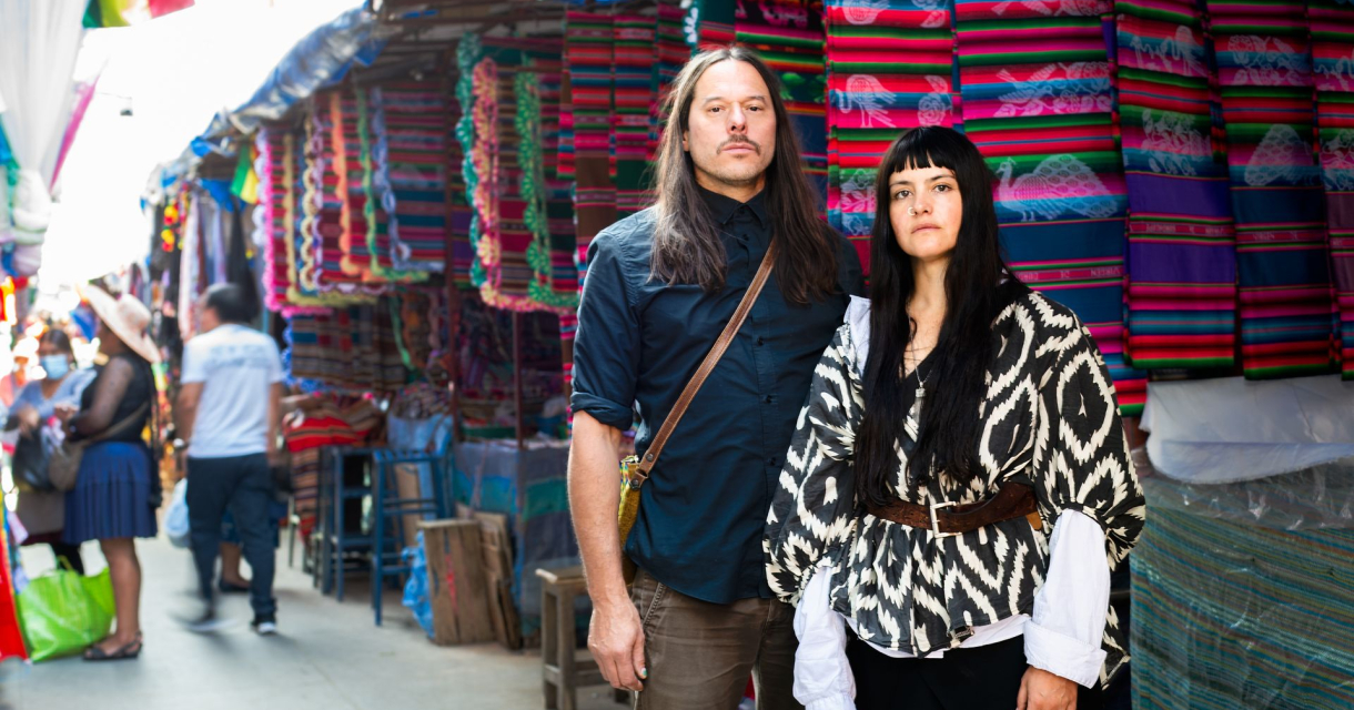 Twee mensen poseren op een markt met felgekleurde doeken en wandkleden aan de kramen om hen heen
