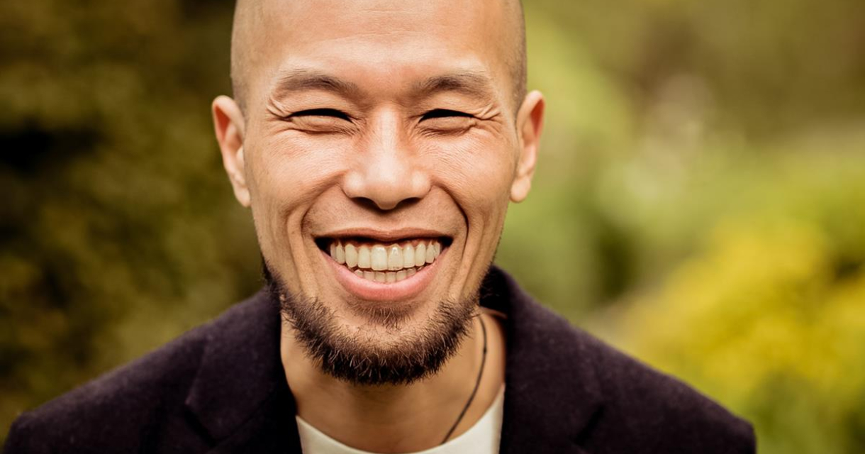 Een man met kort haar en een baard lacht breed in een groene omgeving.