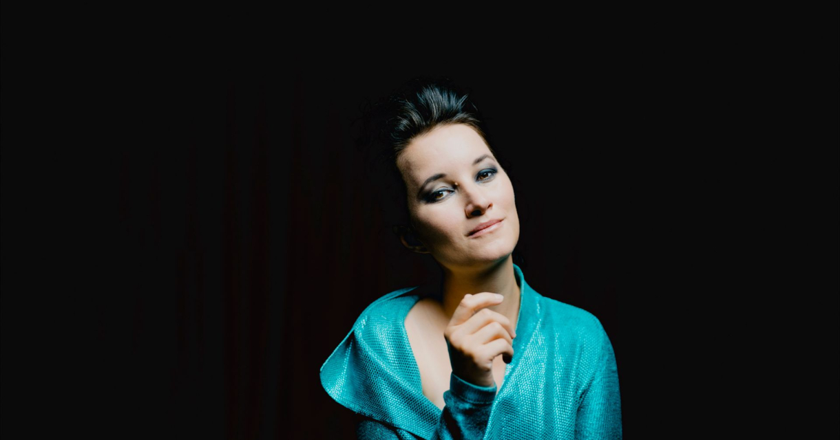 Een vrouw in een turquoise outfit kijkt vriendelijk met een hand onder haar kin.
