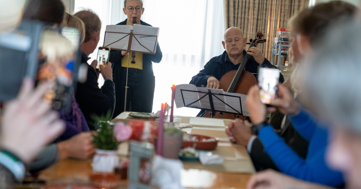 Muzikanten spelen blokfluit en cello voor een groep mensen aan een lange tafel in een huiskamer - fotograaf Umar Chaudry 
