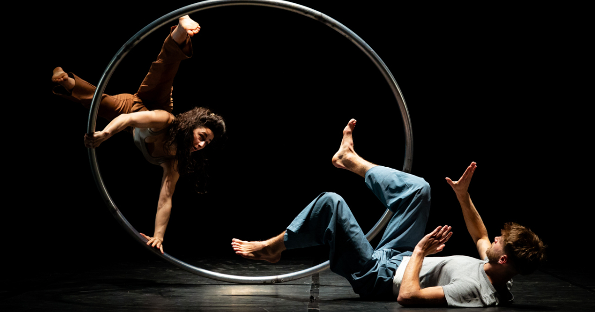 Een acrobate draait zich binnen een metalen ring terwijl een performer op de grond ligt.