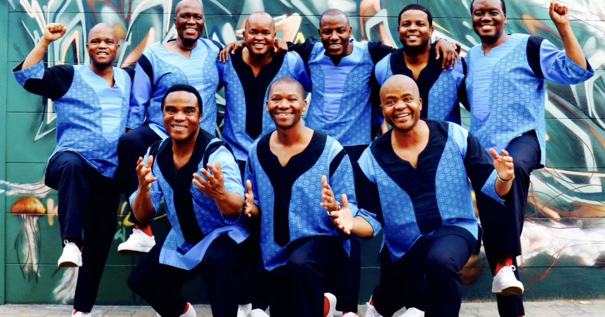 Negen mannen in traditie-inspirerende kleding met blauwe tinten poseren vrolijk voor een muurschildering.