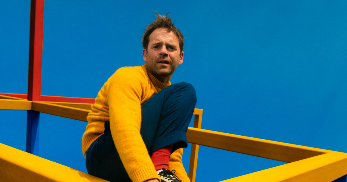 Een man zit op een gele structuur met een blauwe achtergrond.