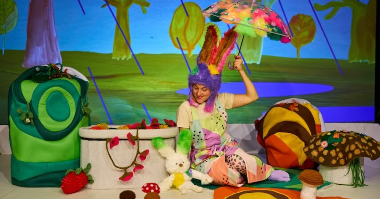 Een vrouw in een kleurrijk kostuum met konijnenoren, zittend in een decor met groenten en fruit.