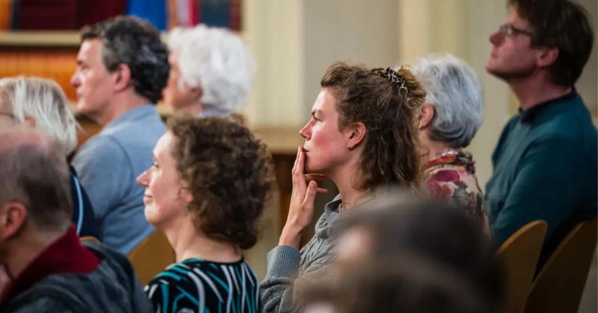 Mensen zittend in een zaal die aandachtig naar iets of iemand luistern. 