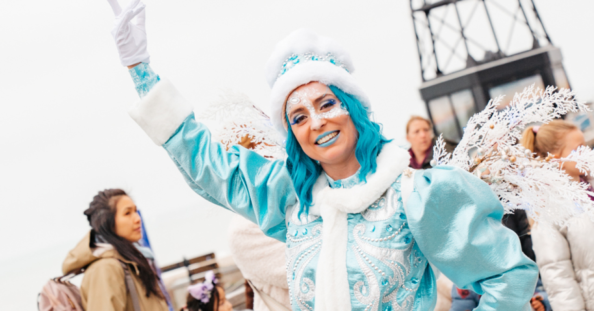 Een performer in een lichtblauwe outfit met vleugels, lachend en de hand omhoog stekend, met een vuurtoren op de achtergrond.