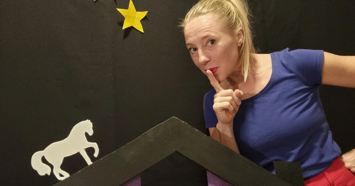 Vrouw in blauwe top legt vinger op haar lip, naast decor met sterren en wit paard op zwart doek