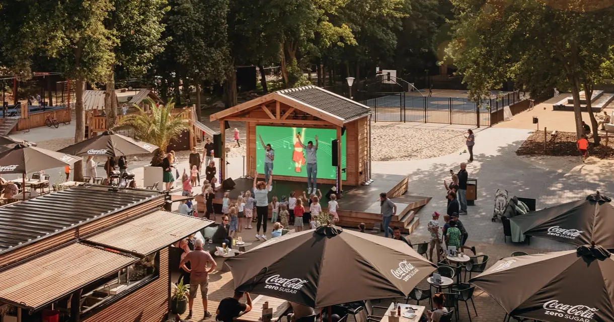 Ein belebter Freizeitbereich mit Menschen, Sonnenschirmen und einem Bildschirm.