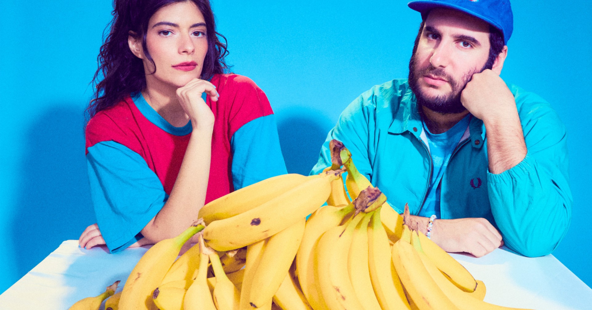 Man en vrouw tegen geen blauwe achterwand aan een tafel vol bananen. 