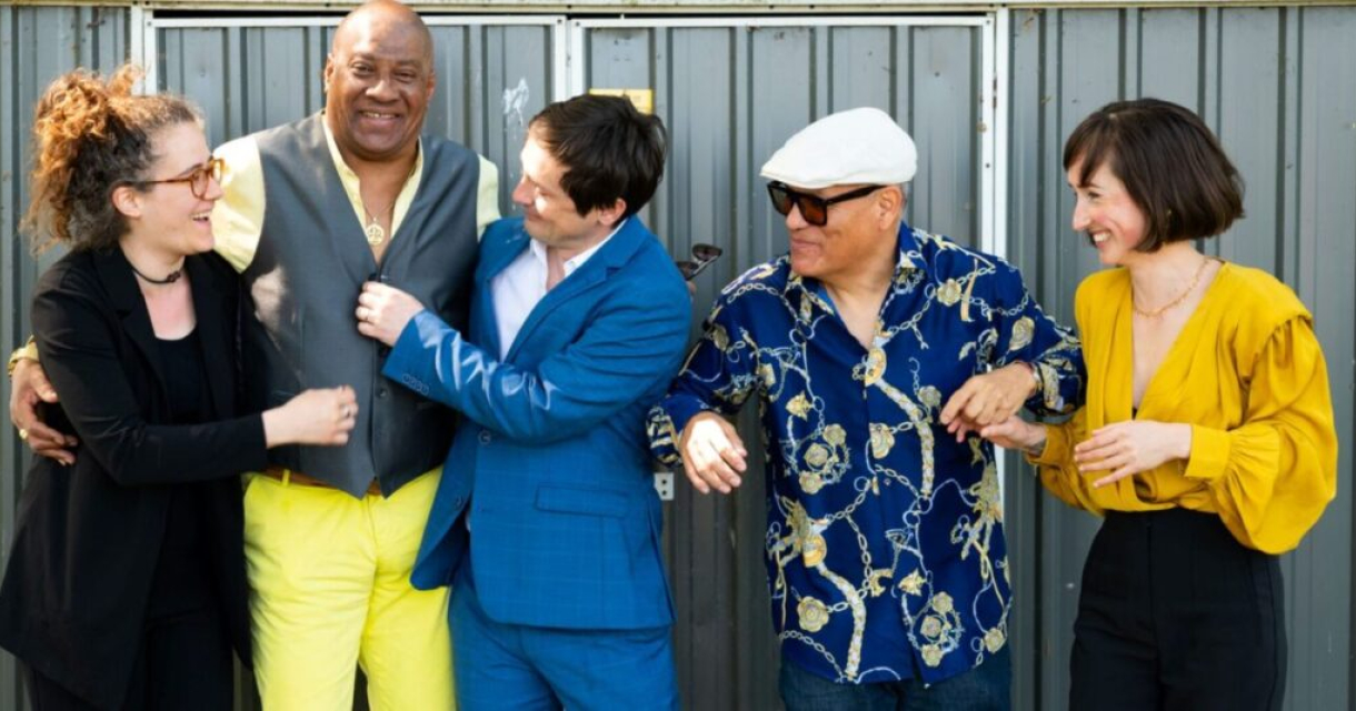 Vijf mensen lachen en poseren samen in een casual setting.