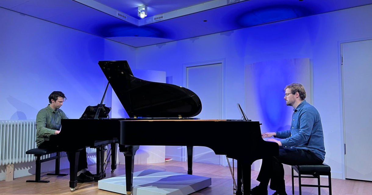 Twee pianisten spelen op vleugels tegenover elkaar in een blauwe verlichte zaal met houten vloer