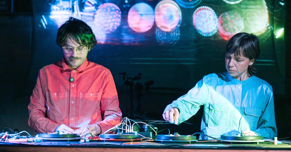 Twee dj’s in felgekleurde outfits mixen live op draaitafels met visuele projecties op de achtergrond