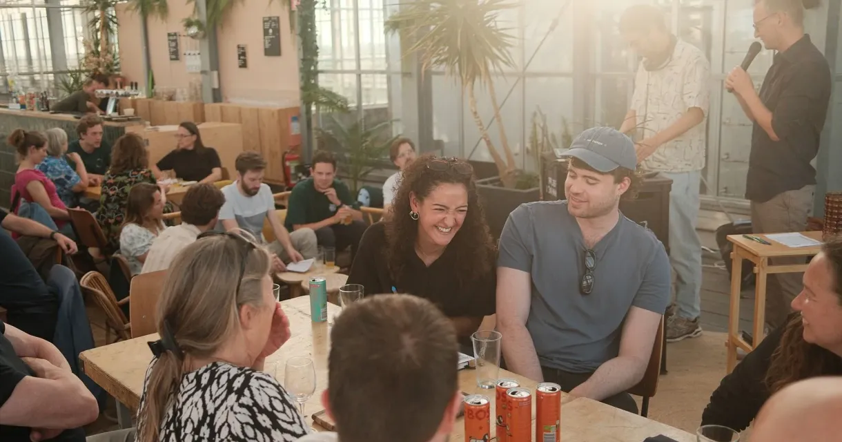 Groep mensen zit aan tafel in een lichte ruimte met planten, lachen en praten.