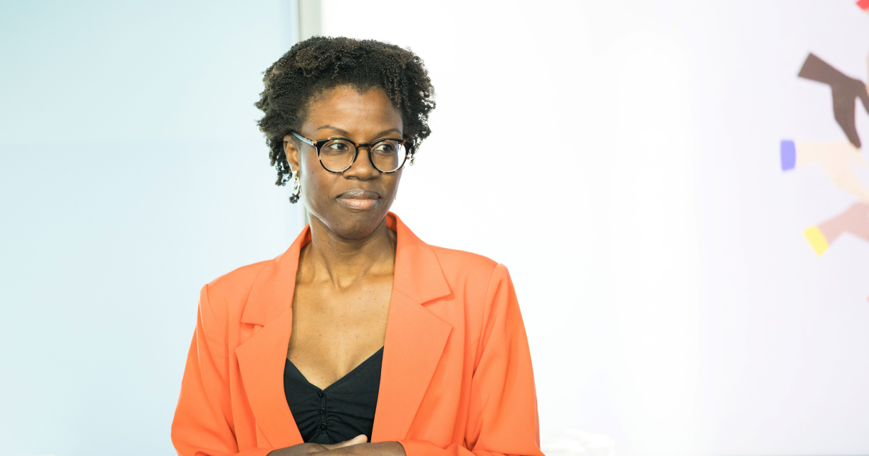 Een vrouw in een oranje blazer spreekt met een serieuze houding.