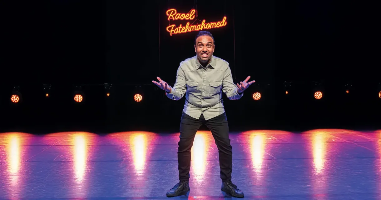 Een stand-up comedian met een grijze blouse, op een podium met lichten, lacht en maakt gebaren.