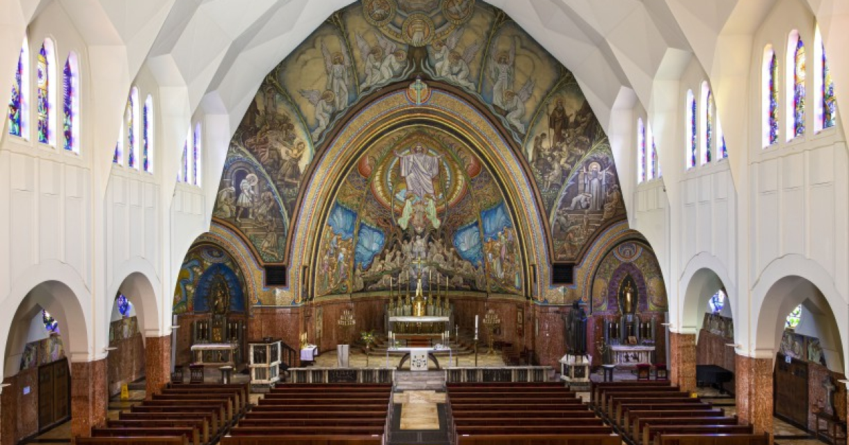 Interieur van een kerk met houten banken, kleurrijke mozaïeken en een groot altaar in het midden