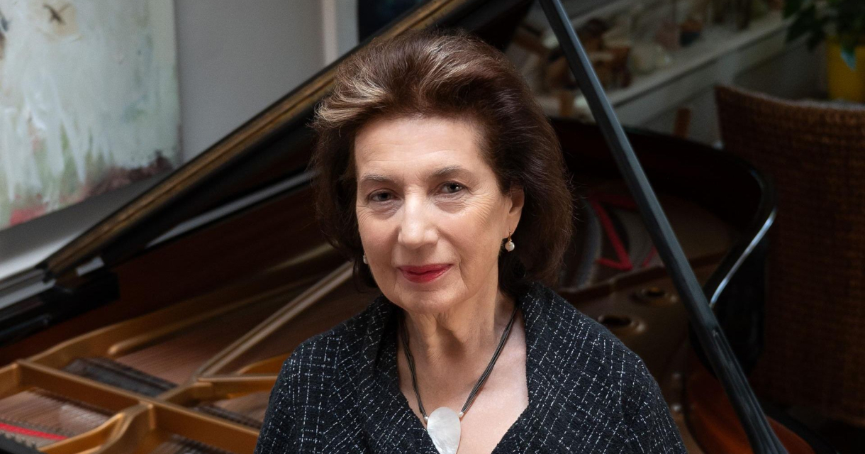 Een vrouw met een donkere blazer en sieraden, staande voor een piano.