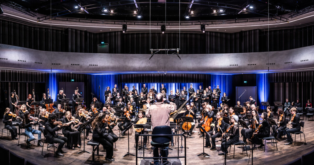 Orkest in spelend in een concertzaal. We zien de dirigent op zijn rug.