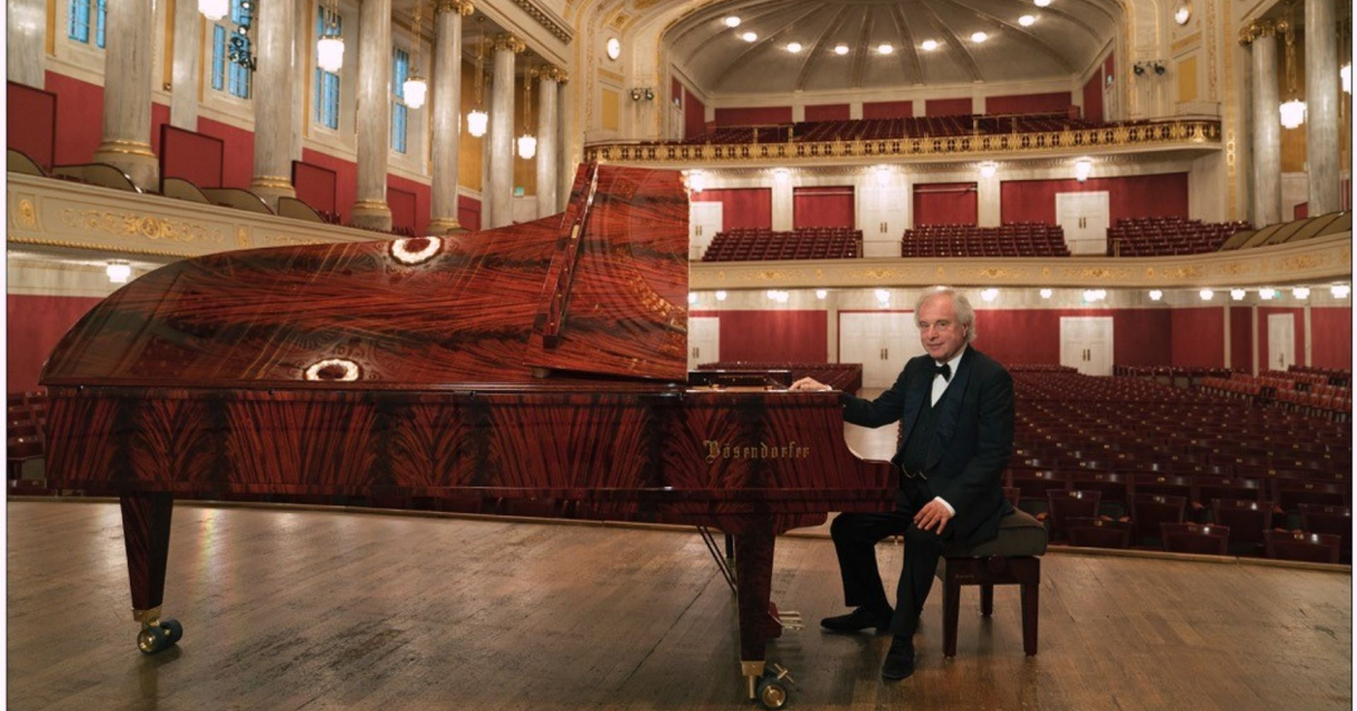 Een pianist in smoking zit naast een fraaie Bösendorfer vleugel op een leeg podium.