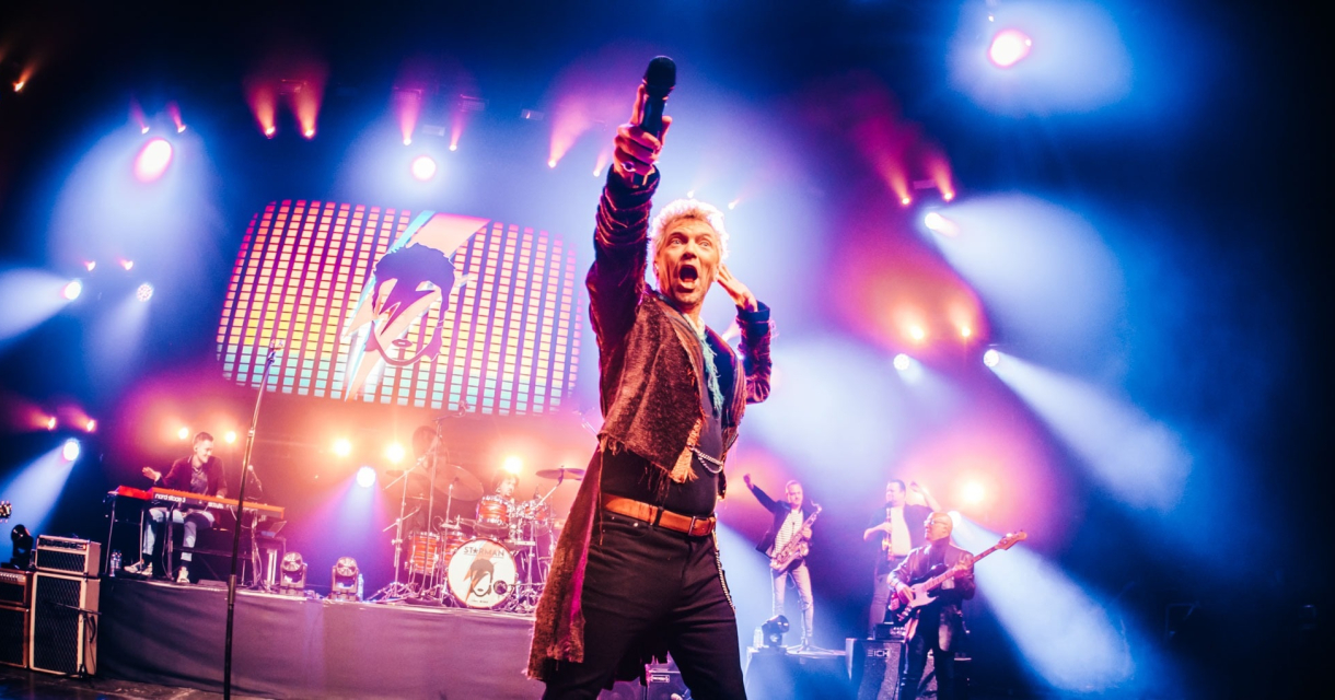A man on stage with a microphone, surrounded by band members and colorful lights.