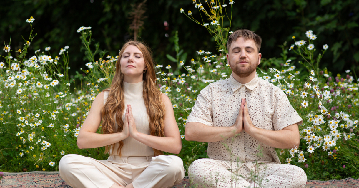 Twee mensen mediteren met samengevoegde handen in een bloemenveld.