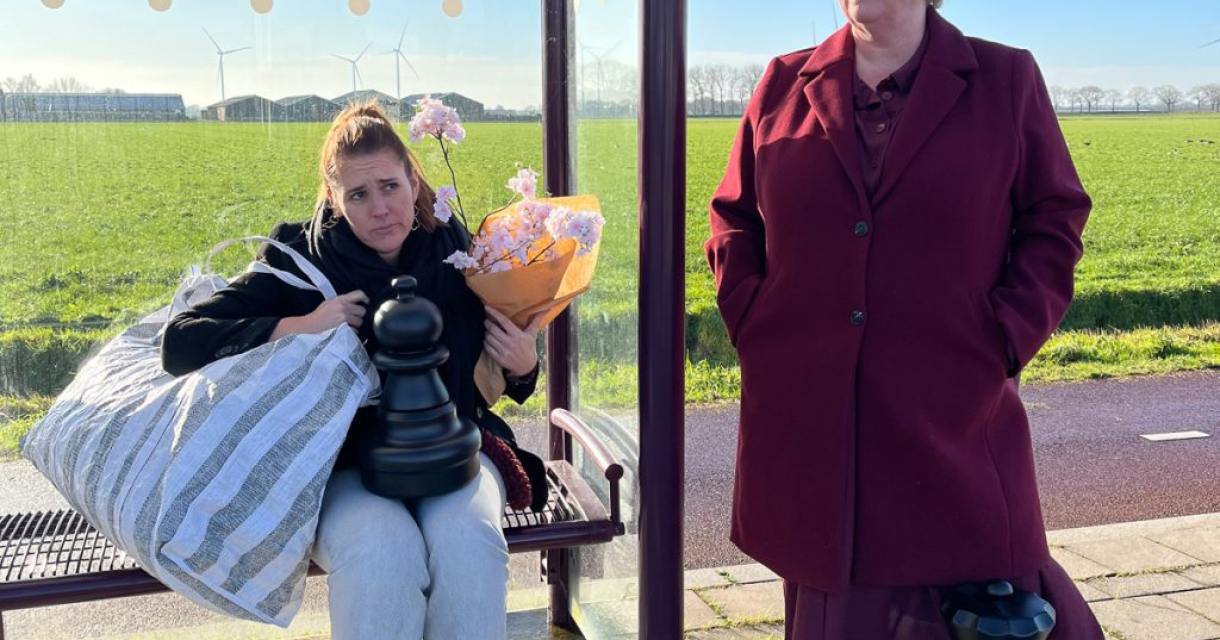 Twee vrouwen staan bij een bushalte, de ene met bloemen.