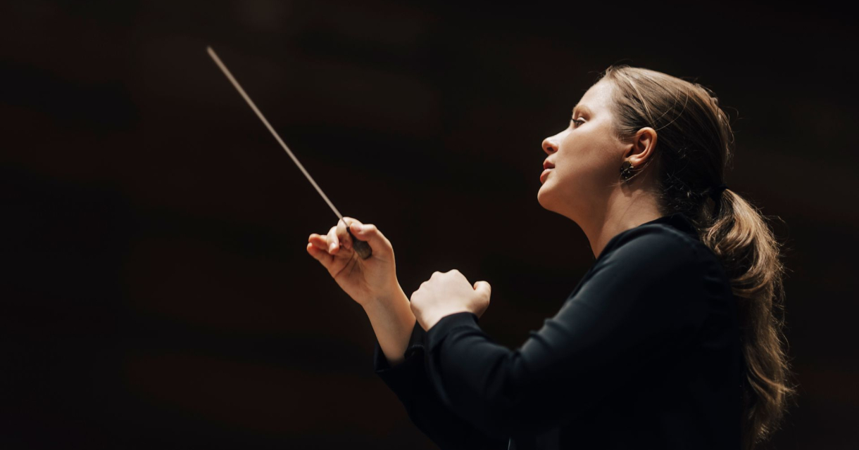 Een vrouwelijke dirigent zwaait met een baton terwijl ze een orkest leidt.
