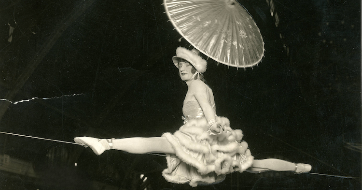 Een acrobate in een witte jurk met een parasol, die op een koord balanceert.