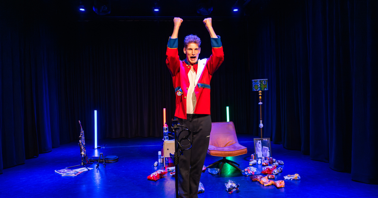 Een performer in een rode jas staat met opgestoken armen op het podium.