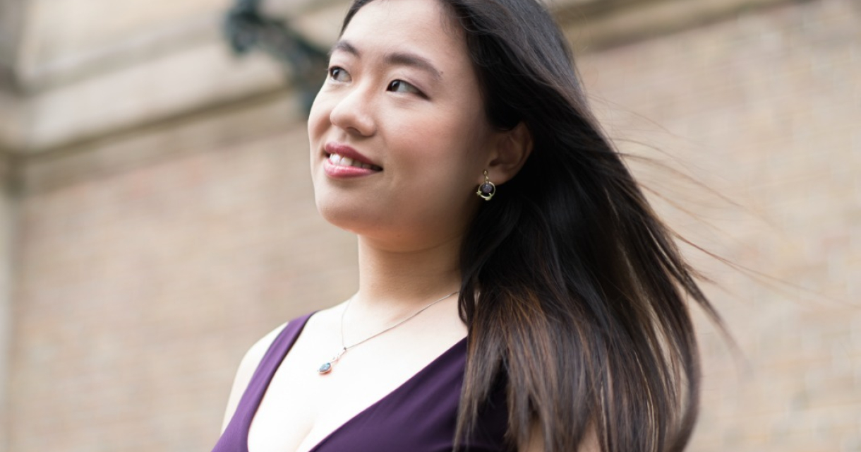 Vrouw met lange haren en een paarse jurk, staand bij een muur.