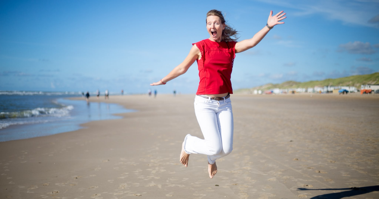 Een enthousiaste vrouw in een rode top springt op het strand in Scheveningen.