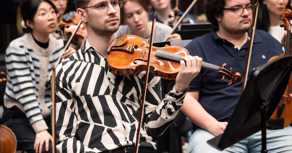 Jongeman met een viool zit in een orkestrepetitie.
