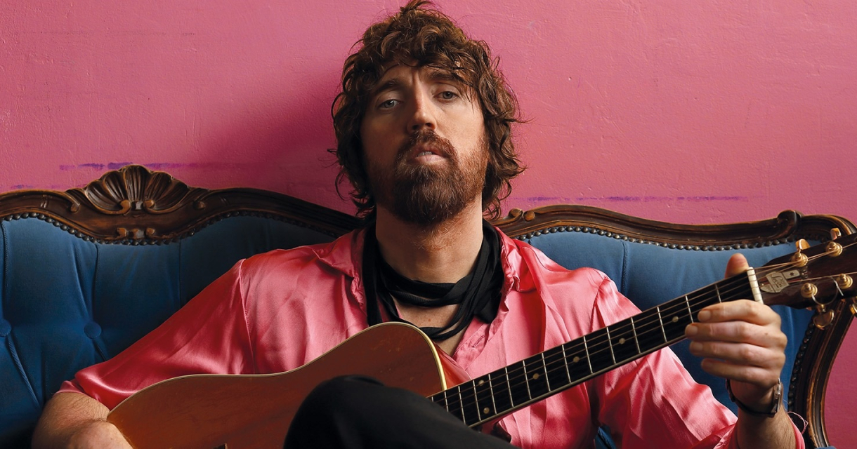 A man in a pink shirt sits on a blue sofa with a guitar.