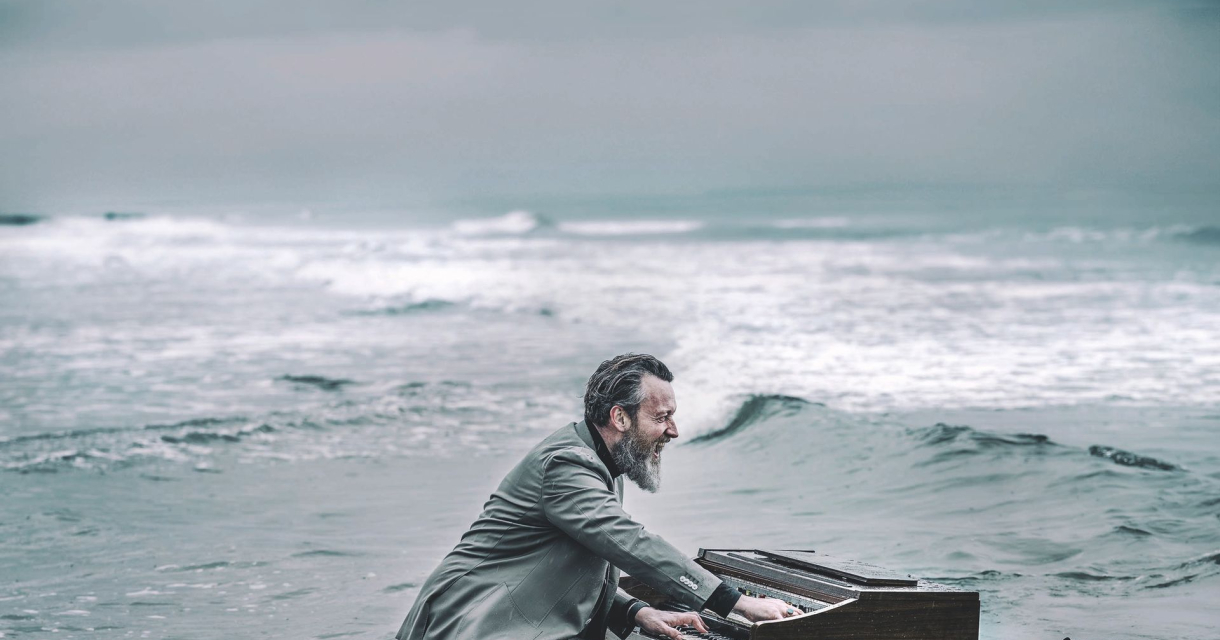 Man in grijs pak speelt piano in ondiep zeewater bij een ruige, bewolkte kustlijn. Man in grijs pak speelt piano in ondiep zeewater bij een ruige, bewolkte kustlijn.