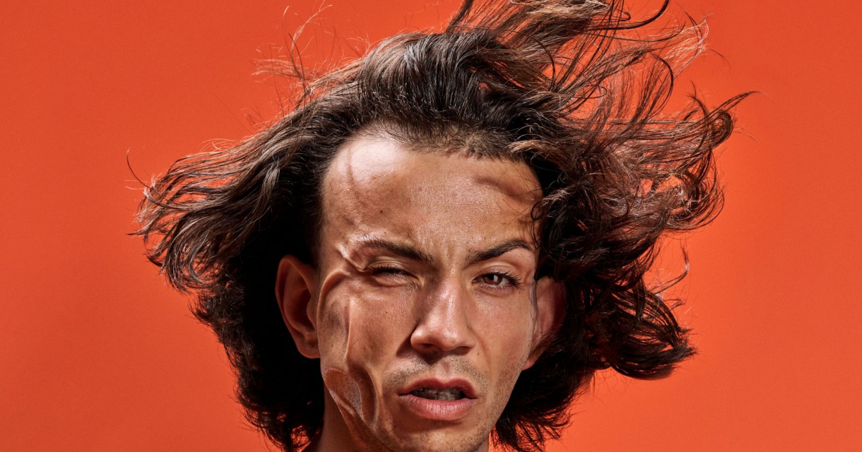 Portret van een man met wapperend haar en verwrongen gezichtsuitdrukking, tegen een feloranje achtergrond. Portret van een man met wapperend haar en verwrongen gezichtsuitdrukking, tegen een feloranje achtergrond.