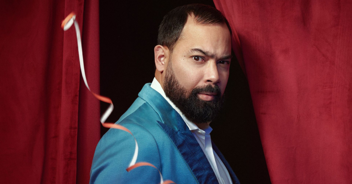 Man die tussen 2 rode gordijnen staat met een turquoise blauw pak aan
