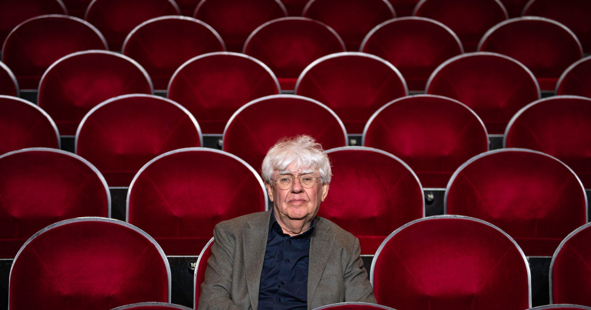 Oudere man met grijs haar zit alleen op een rij in een lege zaal met rode theaterstoelen