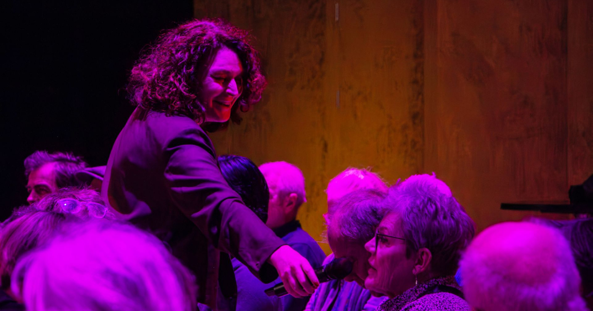 Een presentatrice geeft een vrouw in een roze verlicht publiek het woord door een microfoon aan te geven. 