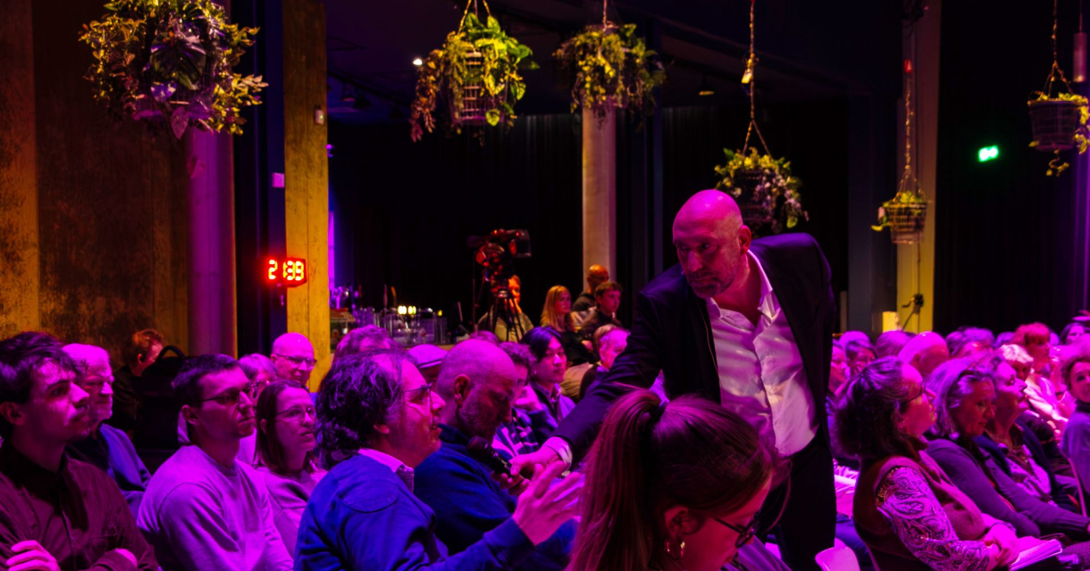 Een presentator in pak geeft een man uit het publiek een microfoon aan. Het publiek zit op stoelen in een roze verlichte zaal 