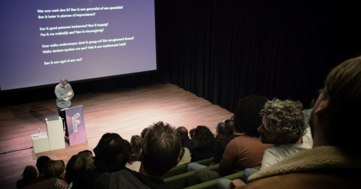 Spreker presenteert voor volle zaal in theater met projectiescherm en donkere gordijnen op de achtergrond Spreker presenteert voor volle zaal in theater met projectiescherm en donkere gordijnen op de achtergrond